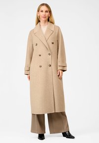 Beige dobbeltbrystet uldfrakke med brede revers, store brune knapper og design der går til knæene, stylet over lysebrune bukser og sorte støvler.