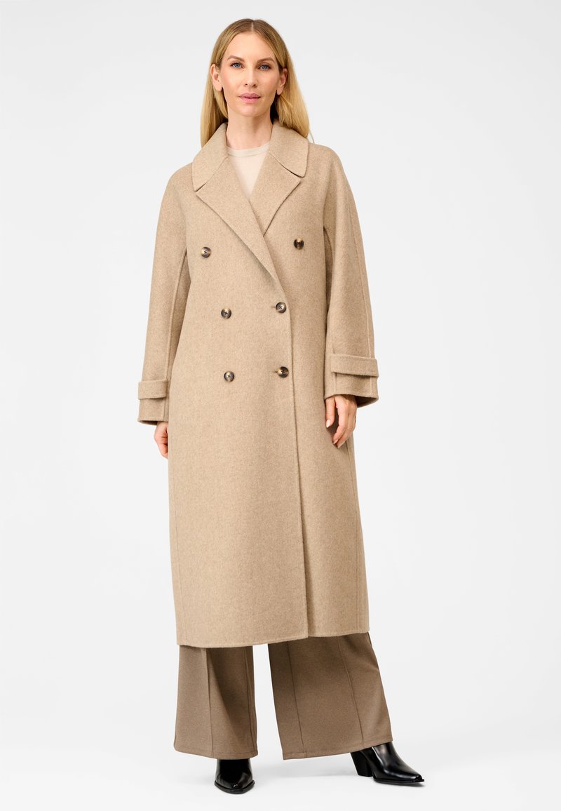 Beige dobbeltbrystet uldfrakke med brede revers, store brune knapper og design der går til knæene, stylet over lysebrune bukser og sorte støvler.
