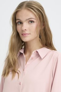 Giovane donna con lunghi capelli biondi, che indossa una camicetta rosa chiaro con bottoni, mentre guarda leggermente di lato con un'espressione neutra.