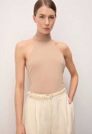 Nő bézs színű, ujjatlan, magas nyakú felsőt visel, ami be van tűrve magas derekú, krémszínű nadrágba, melyen összhangzó öv található, kezét a zsebébe teszi.