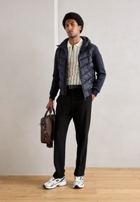 Puffer jacket with a hood, navy color, paired with a striped beige polo, black trousers, and gray sneakers. Brown briefcase in hand.