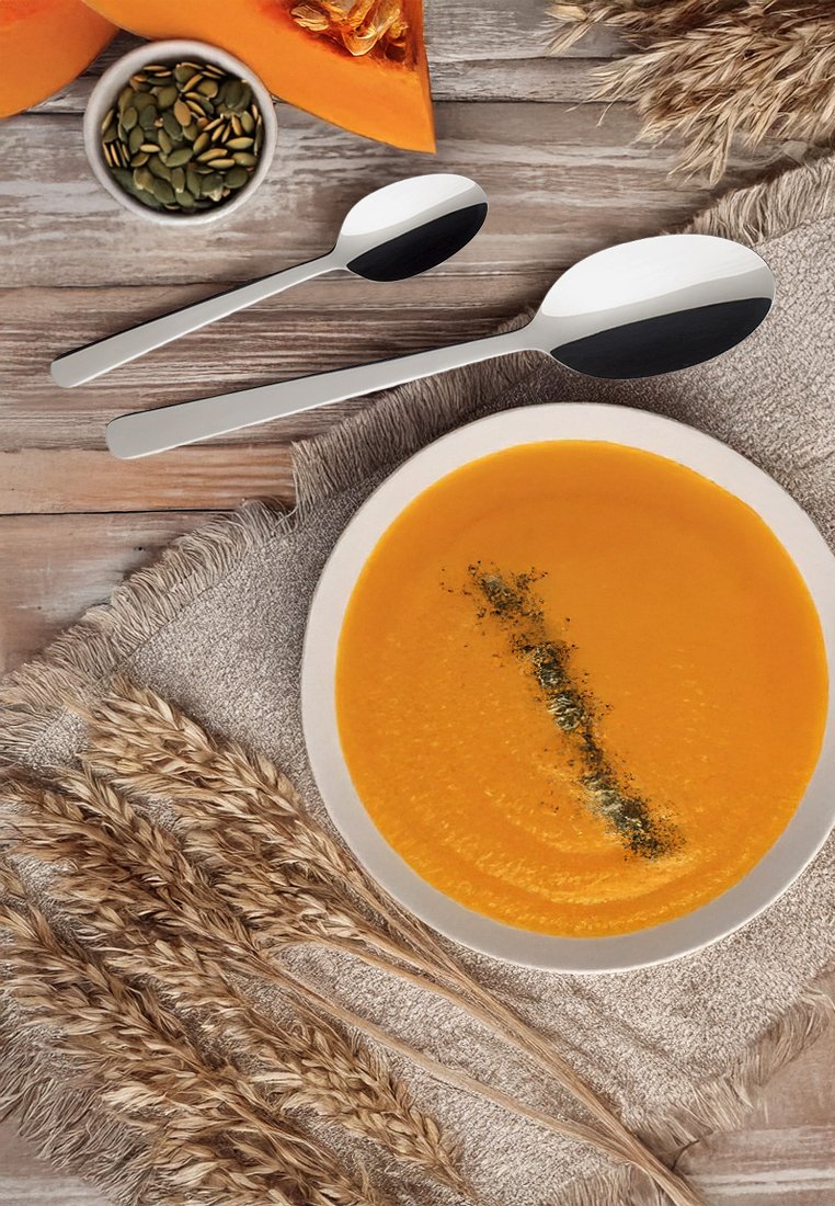 Orange Suppe, garniert mit Kräutern, in einer weißen Schüssel, auf einem strukturierten beigen Tuch platziert. Zwei Edelstahllöffel und Kürbisscheiben in der Nähe.