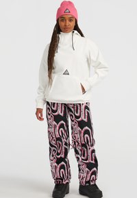 Witte pullover met een voorzak en roze accenten, gecombineerd met zwarte broek met een draaiend roze en wit patroon. Zwarte laarzen.