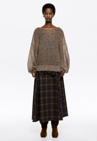 Žena v voľnom hnedom pletenom svetri, s tmavým károvaným dlhým sukňou a hnedými semišovými čižmami, postavená na bielom pozadí.