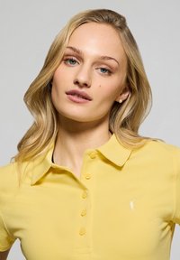 Żółta koszulka polo z teksturowanej tkaniny, z klasycznym kołnierzykiem, guzikami z przodu oraz małym logo na lewej piersi.