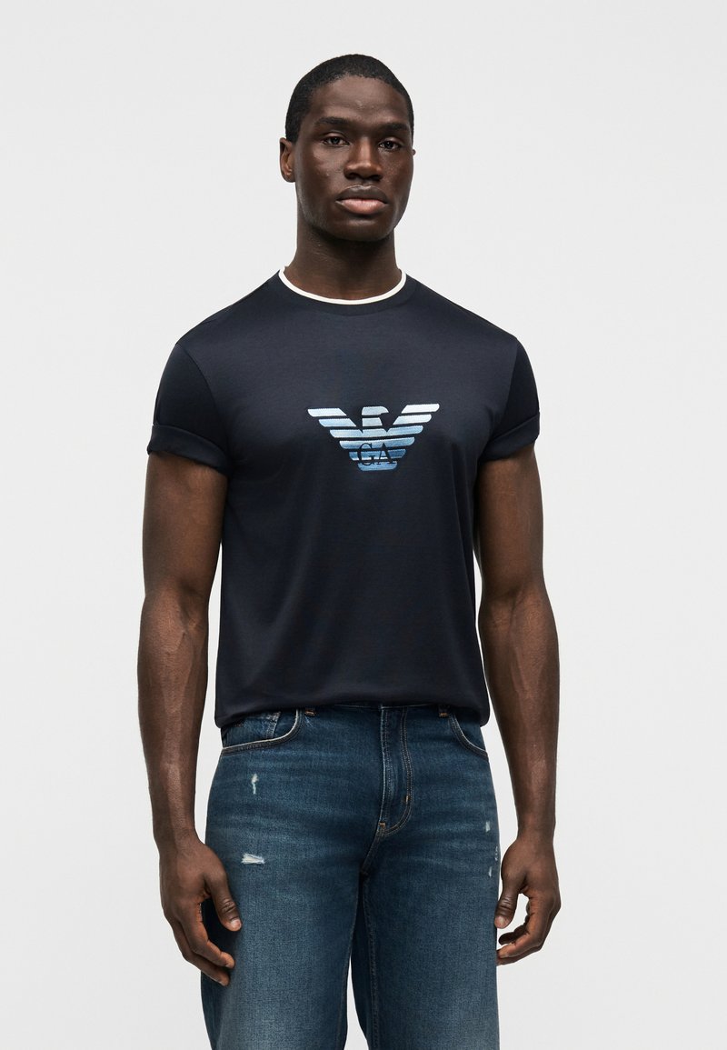 Μπλε σκούρο κοντομάνικο μπλουζάκι με αντίθετο γιακά, διακοσμημένο με βαθμιδωτό λογότυπο αετού σε μπλε και γκρι, σε συνδυασμό με ξεβαμμένο μπλε τζιν.