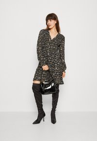 Vestido preto com flores, mangas longas, cintura marcada e saia rodada; combinado com botas de salto alto acima do joelho e uma bolsa preta brilhante.