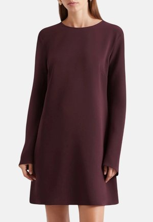 Donna che indossa un semplice vestito color prugna, a maniche lunghe, lungo fino al ginocchio con scollo rotondo su sfondo uniforme.