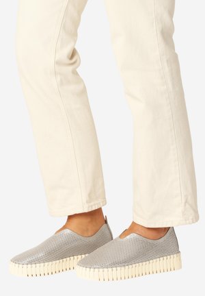 Person iført lyse beige bukser og grå perforerede slip-on sko med tykke cremelignende såler, stående mod en ensfarvet baggrund.