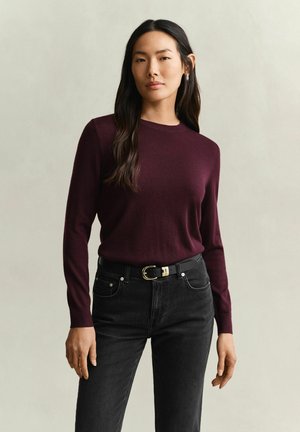 Frau mit langen dunklen Haaren, die einen dunkelvioletten Pullover und schwarze Jeans mit schwarzem Gürtel trägt, steht vor einem einfarbigen hellen Hintergrund.