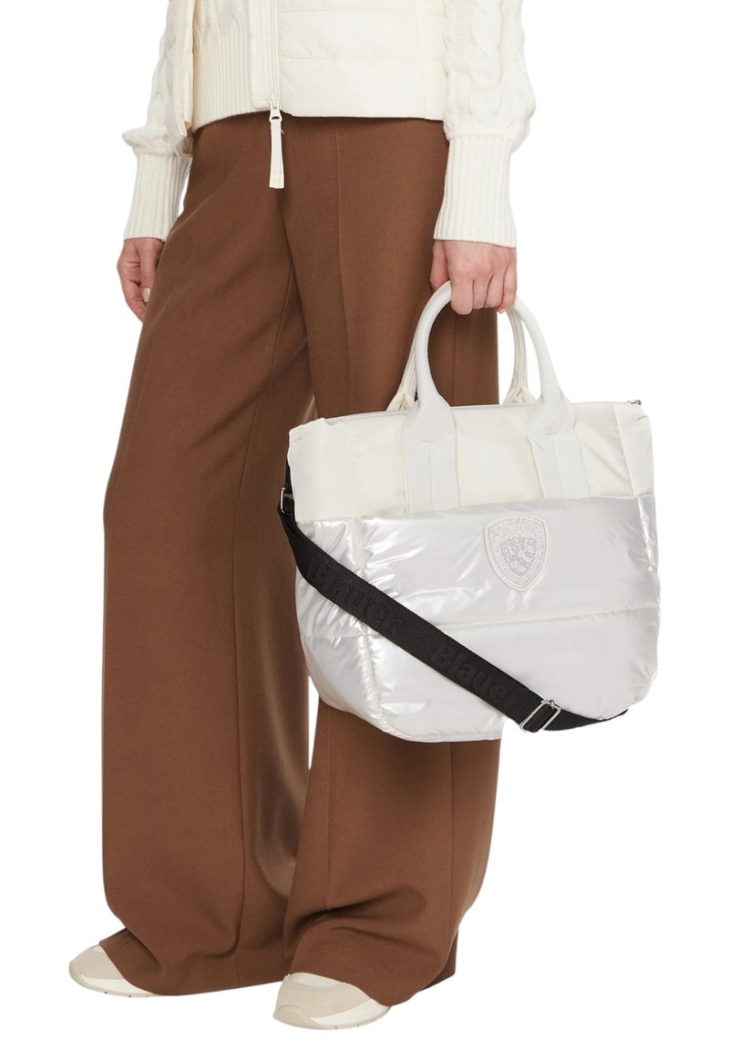 Borsa tote bianca e imbottita con manici superiori e tracolla, caratterizzata da una patch con il logo e una texture lucida, abbinata a pantaloni marroni a gamba larga.