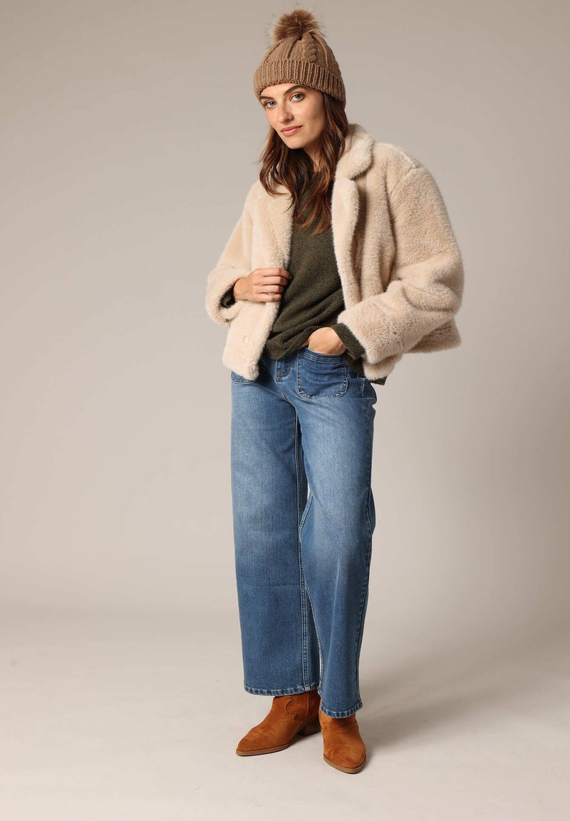 Beige fuzzy jas, olijfgroene trui, lichtblauwe wijde jeans en bruine suède enkellaarsjes, afgemaakt met een gebreide bruine beanie.