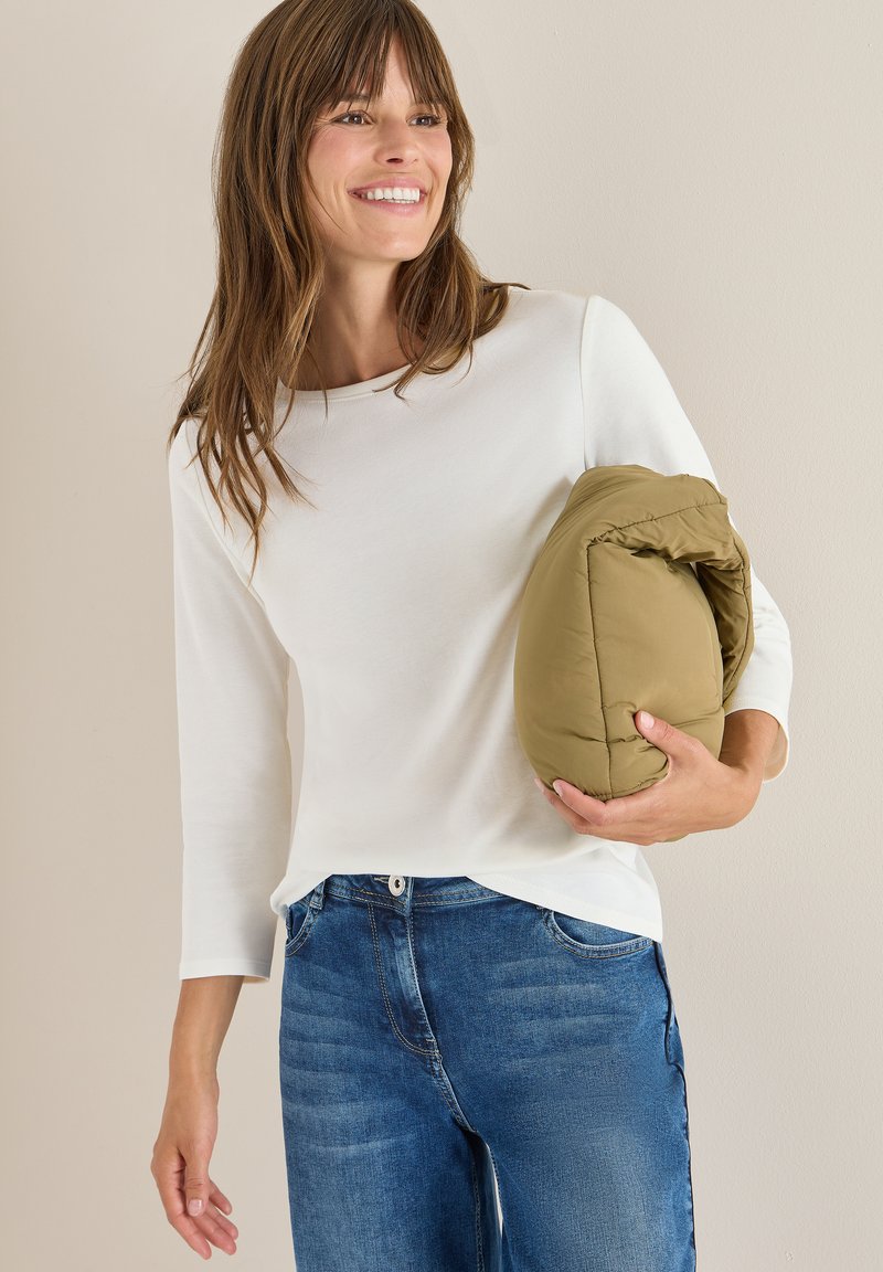 Witte lange mouwen top gecombineerd met middelblauwe jeans. Het model houdt een opgevouwen, olijfgroene pufferjack vast, wat een casual, gelaagde look laat zien.