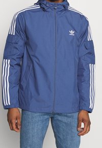 Marinblå jacka med dragkedja fram, huva och vita tre-rands detaljer på ärmarna. Har två sidofickor och elastiska muddar.