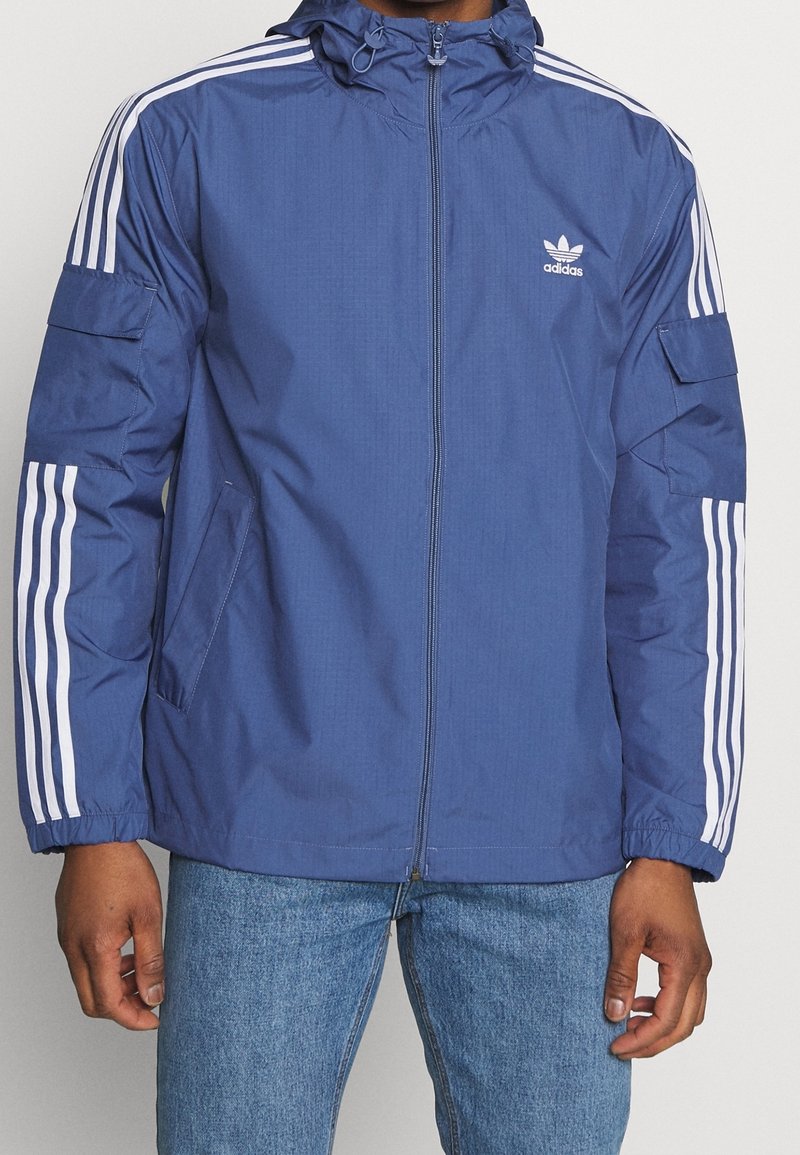 Marinblå jacka med dragkedja fram, huva och vita tre-rands detaljer på ärmarna. Har två sidofickor och elastiska muddar.