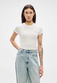 Ung kvinde med langt mørkt hår iført en tætsiddende hvid t-shirt og lyseblå højtaljede jeans, som viser tatoveringer på sin højre arm.