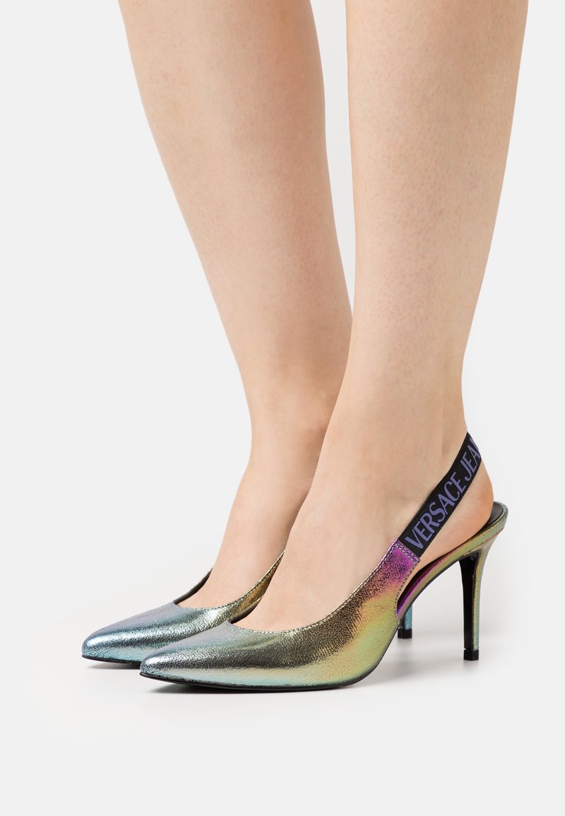 Jambes portant des escarpins slingback métalliques à bout pointu avec des brides élastiques noires estampillées "Versace Jeans".