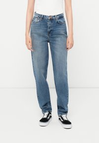 Ανοιχτό μπλε τζιν με ίσια γραμμή και ψηλή μέση, υφή τζιν, πέντε τσέπες και αντίθετη ραφή, συνδυασμένα με μαύρα αθλητικά παπούτσια.