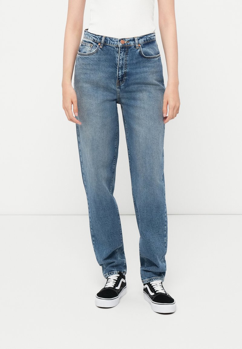 Ανοιχτό μπλε τζιν με ίσια γραμμή και ψηλή μέση, υφή τζιν, πέντε τσέπες και αντίθετη ραφή, συνδυασμένα με μαύρα αθλητικά παπούτσια.