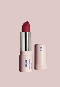 KORRES Barra de Labios Verdadera de Terciopelo en un tubo rosa suave con texto azul, presenta una bala de barra de labios roja profunda y redondeada con una tapa metálica plateada.
