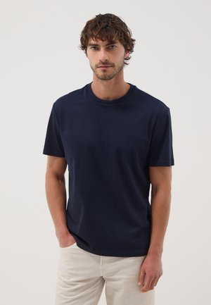 Hombre con pelo rizado y castaño que lleva una camiseta de manga corta azul marino y pantalones beige claros con una mano en el bolsillo sobre un fondo liso.