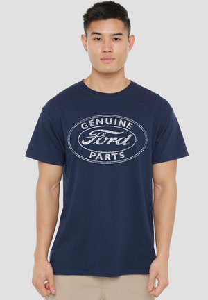 Young man wearing navy blue T-shirt with white "Genuine Ford Parts" logo, standing against plain light gray background.
