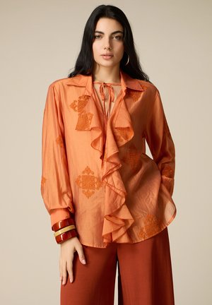 Donna con lunghi capelli scuri indossa una camicetta arancione con volant e motivi ricamati, pantaloni color ruggine e grossi braccialetti a un polso.