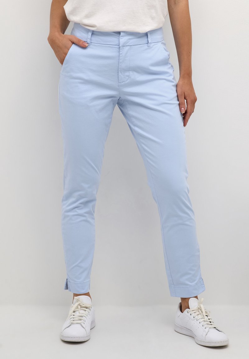 Femme portant un pantalon slim bleu clair et des baskets blanches, une main dans la poche, debout devant un fond uni.