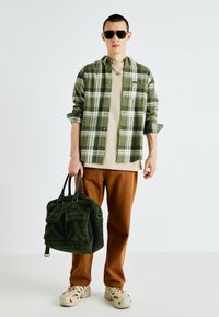 Mężczyzna w zielonej koszuli w kratę, beżowym t-shircie, brązowych spodniach, beżowych sneakersach, w okularach przeciwsłonecznych, trzymający zieloną torbę z fakturą na białym tle.