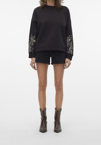 Svart sweatshirt med blommig broderi på ärmarna, ihop med svarta shorts och bruna ankelskängor, med en strukturerad design och textur.