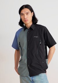 Short-sleeve button-up shirt featuring black, gray, and blue panels, two zippered pockets, and a mountain logo detail. Smooth texture.
