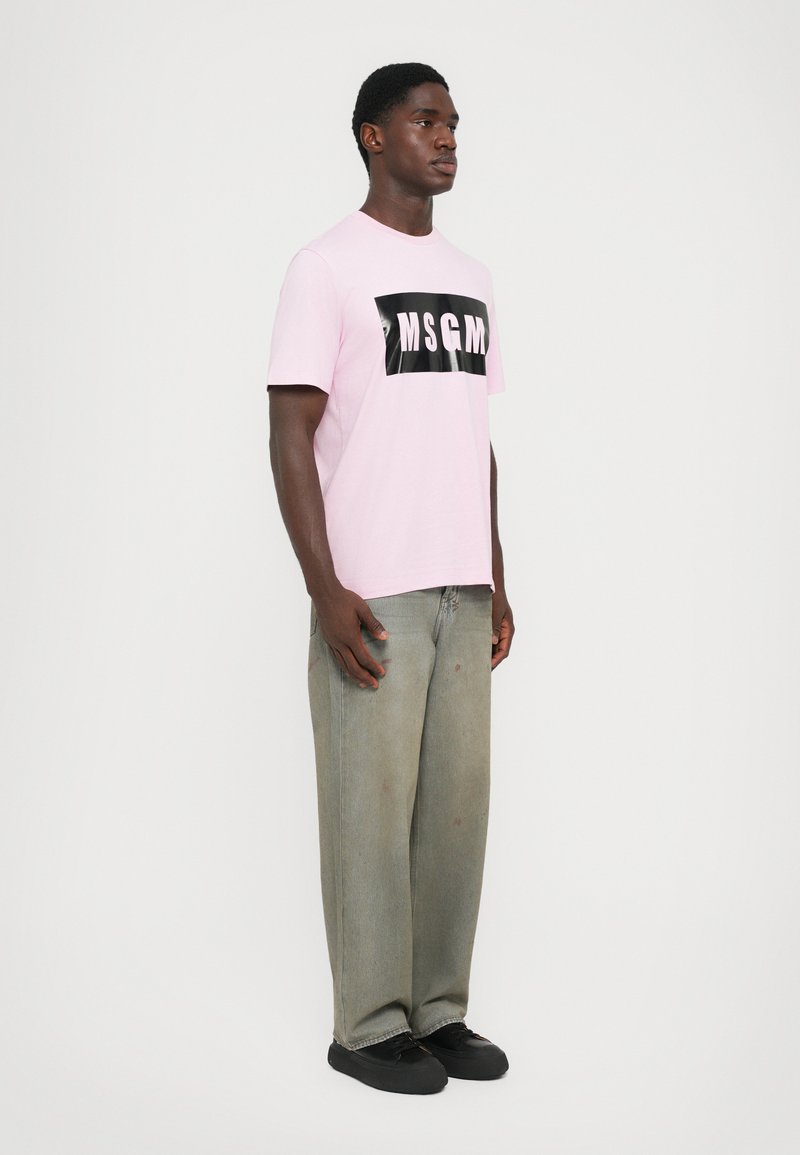 Pink cotton t-shirt featuring a black "MSGM" logo on the front, paired with loose-fitting, faded green jeans and black shoes.