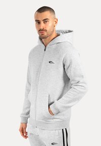 Sudadera gris con cremallera hecha de una tela suave. Cuenta con un bolsillo tipo canguro, puños acanalados y un pequeño logo negro en el pecho.