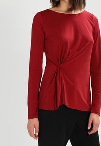 Rotes Langarmoberteil aus weichem Stoff mit einem gedrehten Frontdesign, Rundhalsausschnitt und fließender Form. Kombiniert mit schwarzen Unterteilen.