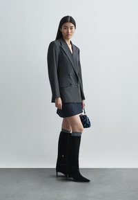 Καπιτονέ γκρι σακάκι με ρίγες, σκούρα τζιν φούστα, μπότες μέχρι το γόνατο από μαύρη σουέντ με μυτερές μύτες, και μικρή ναυτικού χρώματος τσάντα.