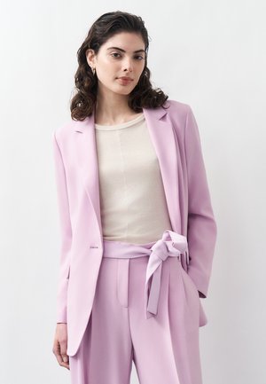 Frau mit welligem dunklem Haar, die ein hellbeiges Oberteil und einen pastellrosa Anzug mit gebundenem Gürtel trägt, vor einem schlichten weißen Hintergrund steht.