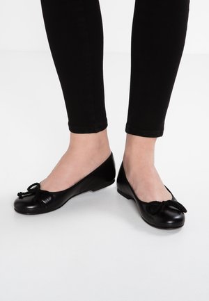 Femme portant des ballerines noires avec des nœuds décoratifs, assorties à un pantalon noir ajusté, sur un fond blanc uni.