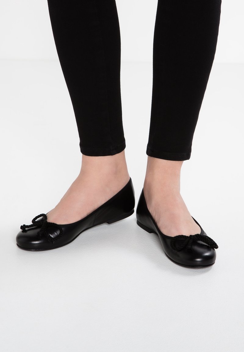 Femme portant des ballerines noires avec des nœuds décoratifs, assorties à un pantalon noir ajusté, sur un fond blanc uni.