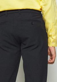 Donkergrijze gestructureerde broek met een subtiel patroon, voorzien van twee achterzakken met knoopsluitingen, gecombineerd met een gele shirt.