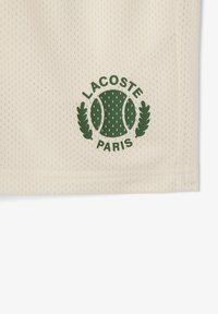 Un tissu en maille couleur crème présentant un logo vert foncé avec une balle de tennis et des feuilles de laurier, avec le texte "LACOSTE" et "PARIS" en dessous.