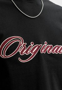 Chemise noire avec le texte "Originals" en cursive bordeaux et blanc, et un collier chaîne argenté autour du cou.