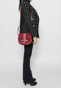 Borsa a tracolla in suede bordeaux con forma arrotondata, dettagli in pelle e chiusura magnetica. Presenta una spalla regolabile e hardware minimal.