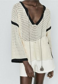Personne portant un pull en crochet crème à col V avec des bordures et des pompons noirs, associé à un short blanc, sur un fond clair et uni.