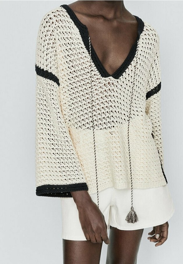 Personne portant un pull en crochet crème à col V avec des bordures et des pompons noirs, associé à un short blanc, sur un fond clair et uni.