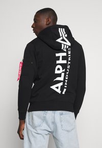 Black hooded sweatshirt with white "ALPHA INDUSTRIES" logo on back, featuring a zip pocket on the sleeve and a red tag accent.