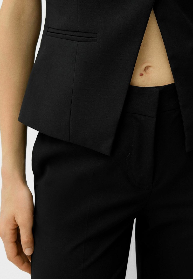 Blazer negro ajustado con un diseño estructurado y una textura sutil, combinado con pantalones negros de tiro alto que cuentan con un bolsillo frontal.