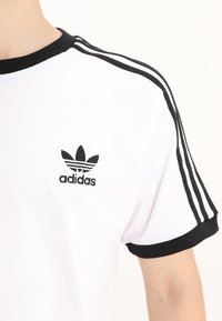 White Adidas t-shirt with black trim on collar and sleeves, featuring black three stripes on shoulder and Adidas trefoil logo on chest.