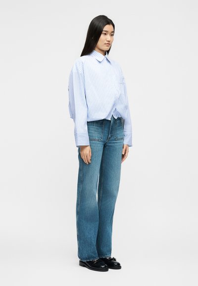 Γυναίκα που στέκεται φορώντας φαρδιά ριγέ πουκάμισο ανοιχτού γαλάζιου χρώματος, ψηλόμεσα ίσια μπλε τζιν και μαύρα μοκασίνια, μπροστά σε λευκό φόντο.