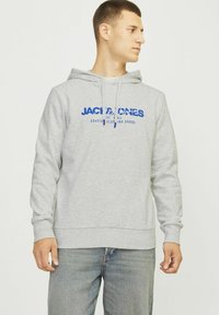 Sudadera gris de material polar, con un bolsillo tipo canguro, capucha con cordón y logo impreso azul en la parte delantera. Combinada con jeans.