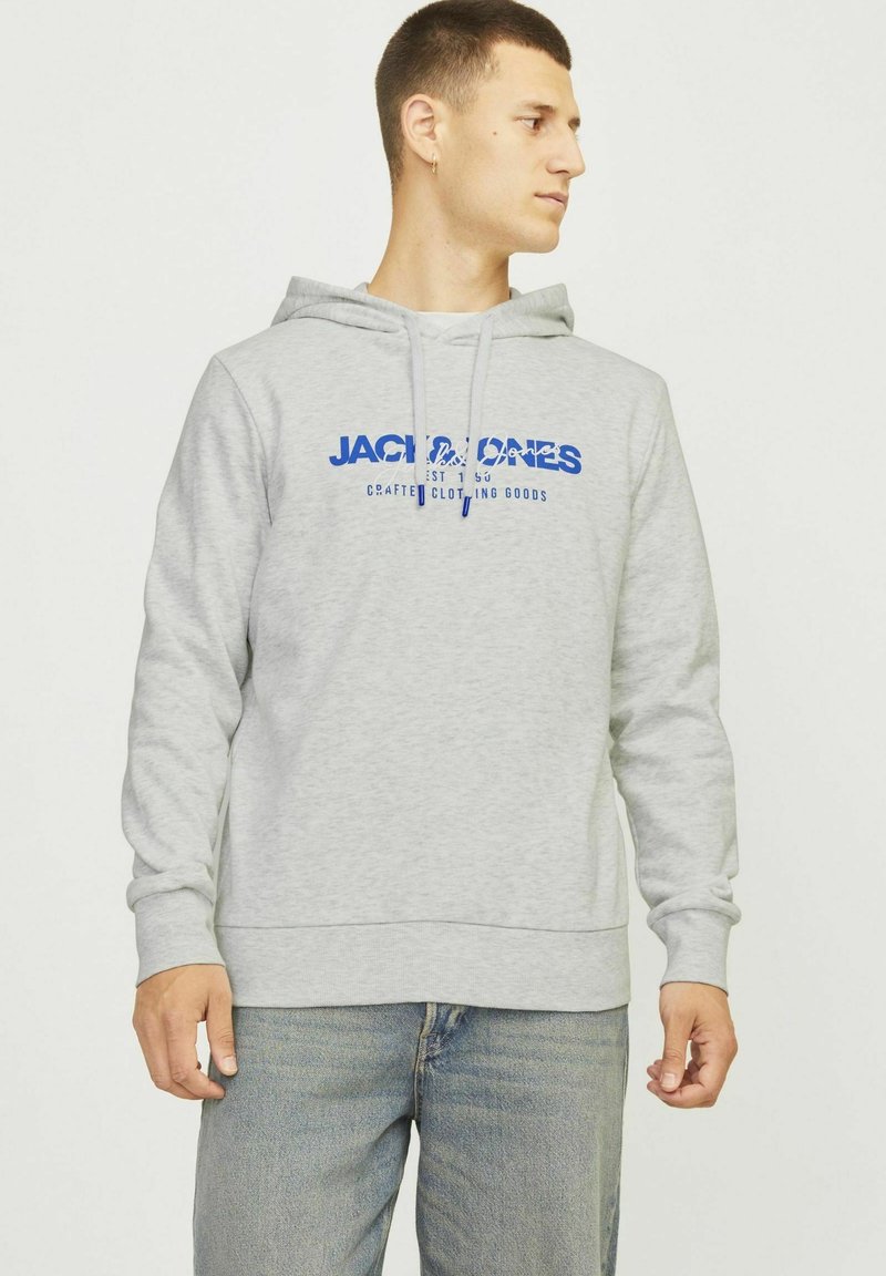 Sudadera gris de material polar, con un bolsillo tipo canguro, capucha con cordón y logo impreso azul en la parte delantera. Combinada con jeans.